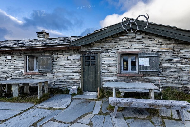 A traditional stone house with a café sign, wooden benches in front, surrounded by a mountainous landscape under a cloudy sky, Peer Gynt hytta, Rondane National Park, Otta, Innlandet, Norway