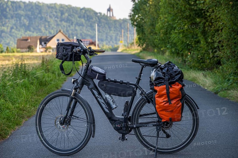An electric bicycle with an orange bag stands on a rural road, surrounded by green nature and a hill with houses, touring bike in front of the Kaiser Wilhelm monument, Holzhausen, Porta-Westfalica, North Rhine-Westphalia