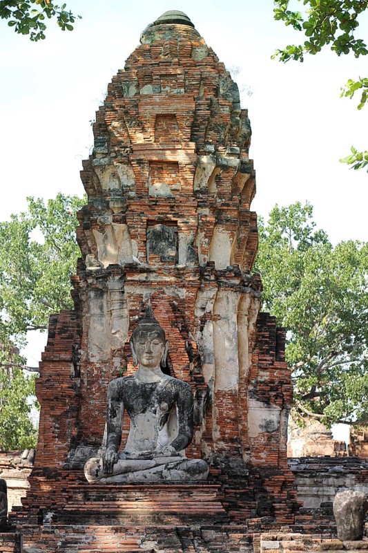 A Buddha statue seated before an ancient brick stupa, surrounded by greenery, exuding serenity in Wat Chaiwatthanaram in Ayutthaya, Thailand