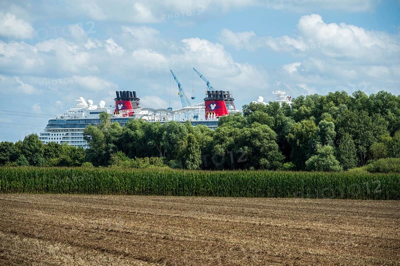 A cruise ship behind trees next to a field under a partly cloudy sky, cruise ship in the shipyard of Papenburg