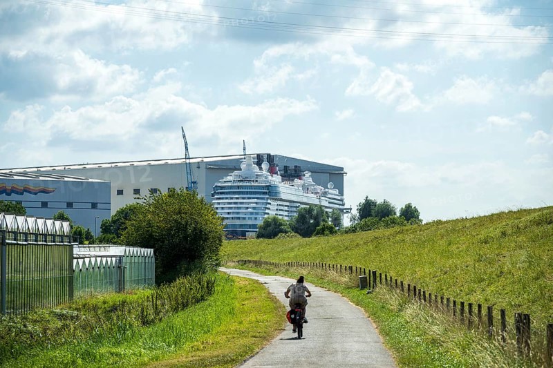 A cyclist on a country road next to a shipyard with a cruise ship in the background, cruise ship at the Meyer shipyard in Papenburg