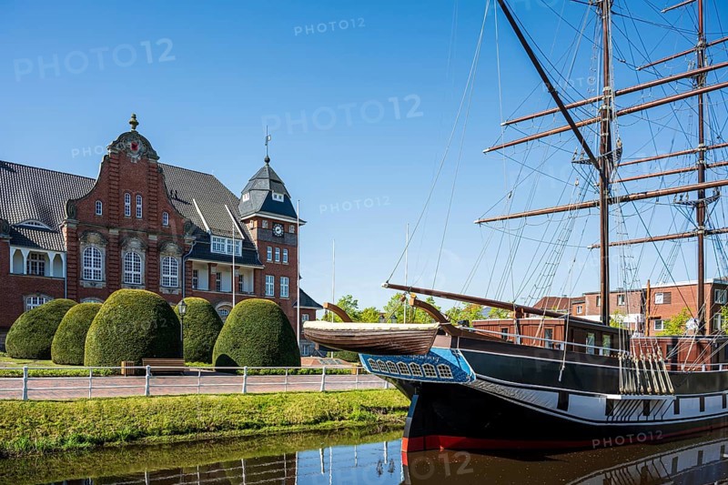 A sailing ship lies in front of a historic building with a blue sky and well-tended bushes on the canal, The sailing ship Frederike von Papenburg in front of the town hall in Papenburg