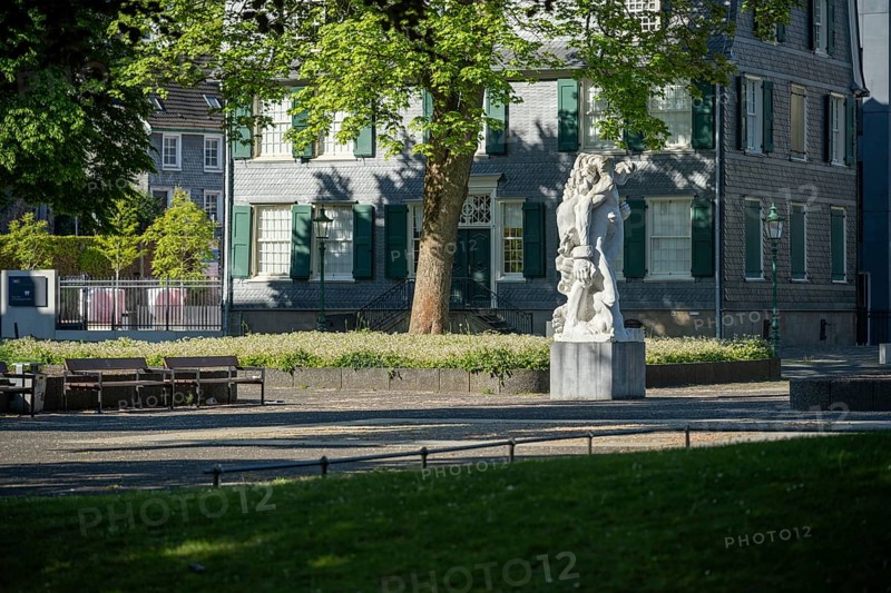 Small square with stone statue in front of an old house with green shutters, illuminated by sunlight, statue The Strong Left by Alfred Hrdlicka in front of the Friedrich Engels House in Wuppertal Barmen