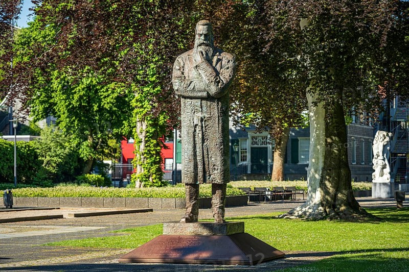 Stone statue of a man with crossed arms in an urban park with surrounding buildings, statue of Friedrich Engels by sculptor Zeng Chenggang in the Engelsgarten Wuppertal Barmen