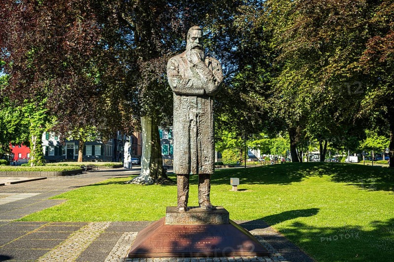 Stone statue of a man with crossed arms in a sunny park with many trees, statue of Friedrich Engels by sculptor Zeng Chenggang in the Engelsgarten Wuppertal Barmen