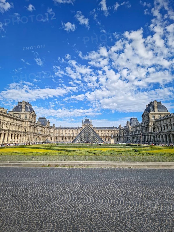 A grand panoramic view of the Louvre Palace with its iconic glass pyramid in the center under a magnificent blue sky with fluffy white clouds in Paris, France