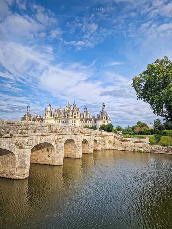 A picturesque stone bridge spans the still water leading to the magnificent Chateau de Chambord. The iconic French Renaissance castle stands under a beautiful sky with soft clouds