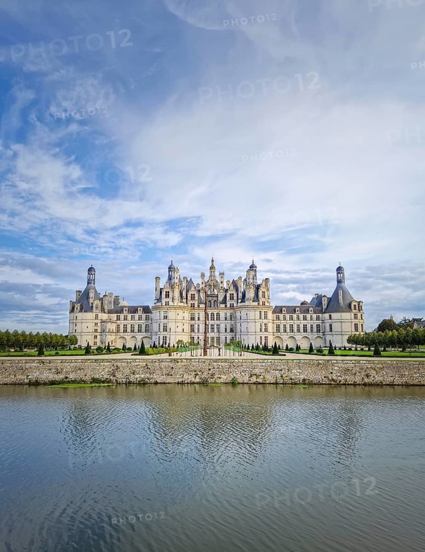 The symmetrical Château de Chambord is reflected in the still water, highlighting its magnificent Renaissance architecture with numerous turrets. The majestic castle stands beneath a beautiful sky