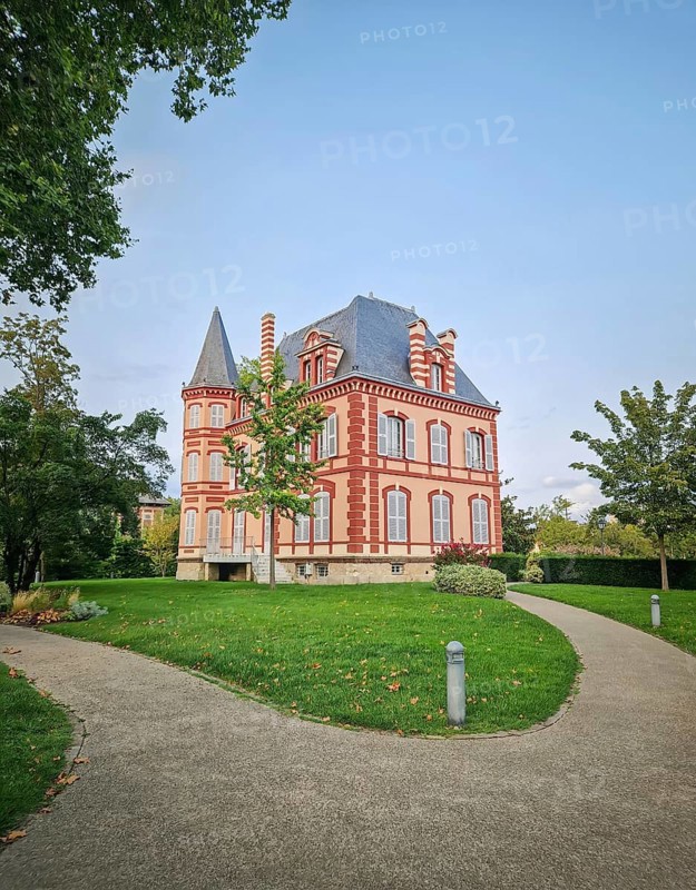 The Castel Rose in L'Isle-Adam, France. Historic mansion characterized by its pink walls and distinctive red brick trim. The building is surrounded by a lush green lawn and winding paths