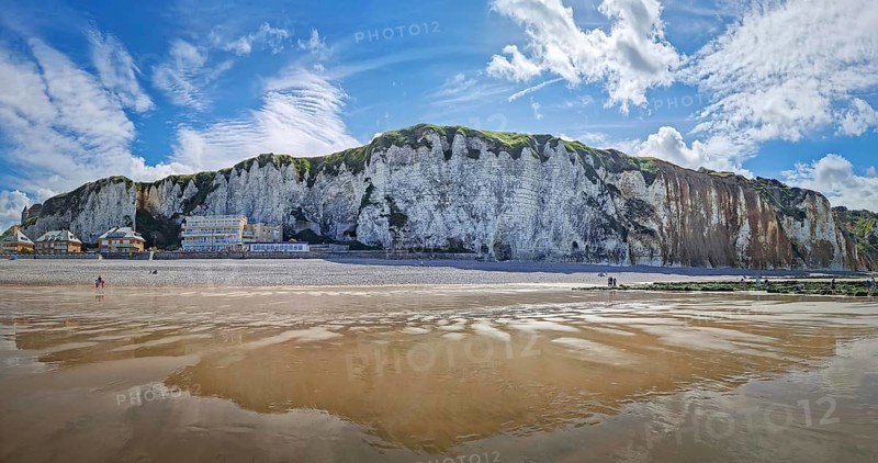 Panoramic view of Dieppe beach in Normandy, France. Majestic white chalk cliffs rise above a pebble shore, with the low tide revealing a vast expanse of wet sand reflecting the blue sky