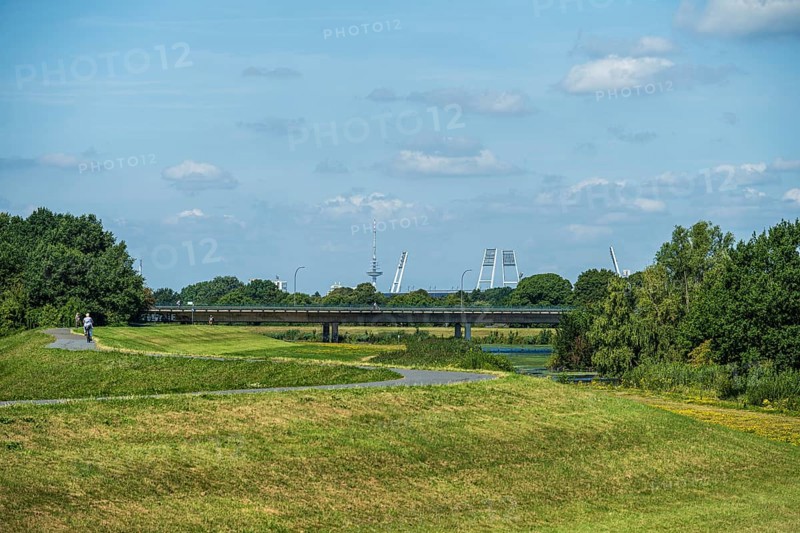 A bridge leads over green areas with trees and a path under a blue sky with few clouds. Stadium towers are visible, Weser cycle path, Weser, floodlight masts Weserstadion, Bremen