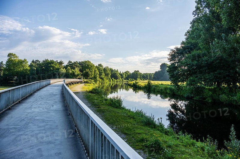 Path along a calm river, lush vegetation and cloudy sky are reflected in the water, Emssee near Warendorf, Münsterland