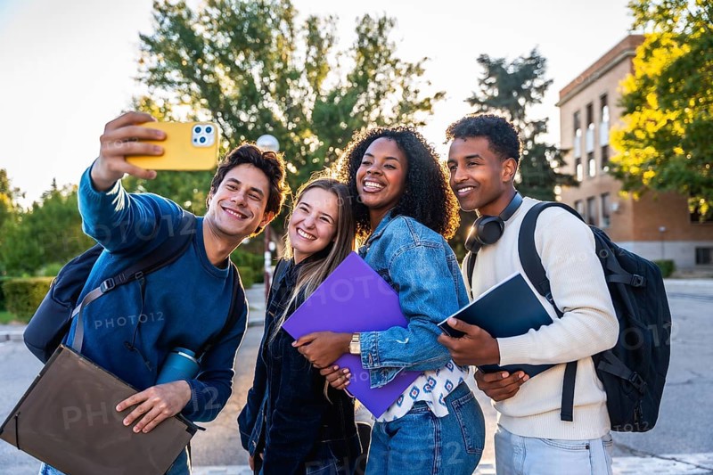 Group of smiling multi ethnic students standing outdoors on campus, celebrating friendship, youth, and education while taking a cheerful selfie together in bright sunlight