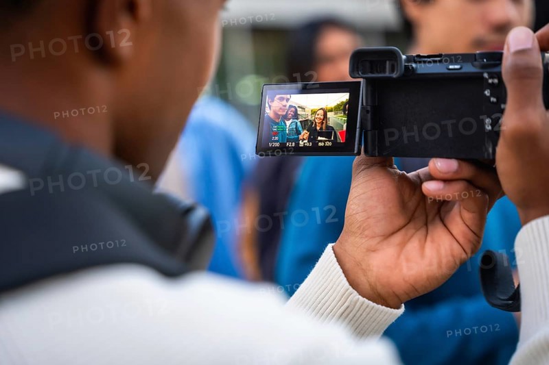 Young person holding a digital camera, filming three diverse students smiling and engaging in conversation on a university campus, capturing modern education and youth content creation