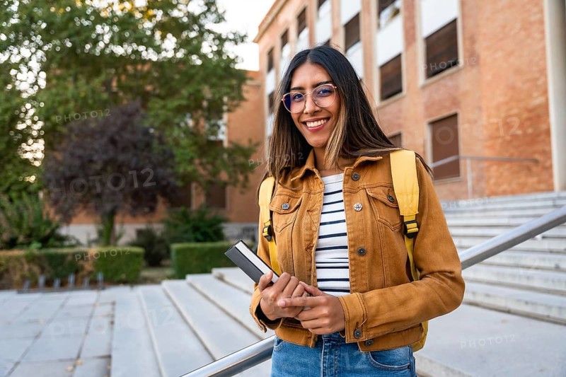 Young woman smiling, wearing glasses and a backpack, holding a book, and standing on university stairs, representing education, learning, and future success