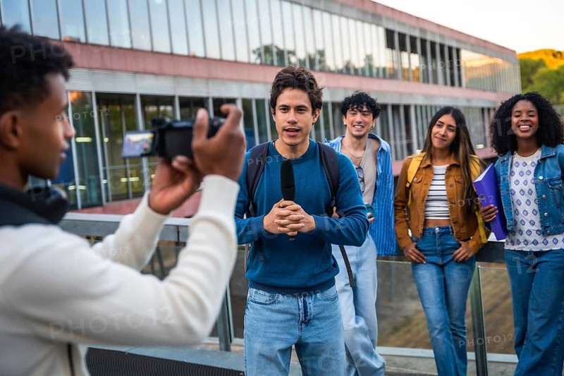 Group of diverse university students collaborating, with one student holding a microphone and speaking into a camera being filmed by another, creating media content on university grounds