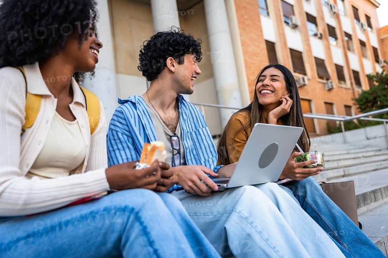 Group of diverse university students sharing a happy moment, laughing and eating lunch together on campus stairs, symbolizing friendship and student life