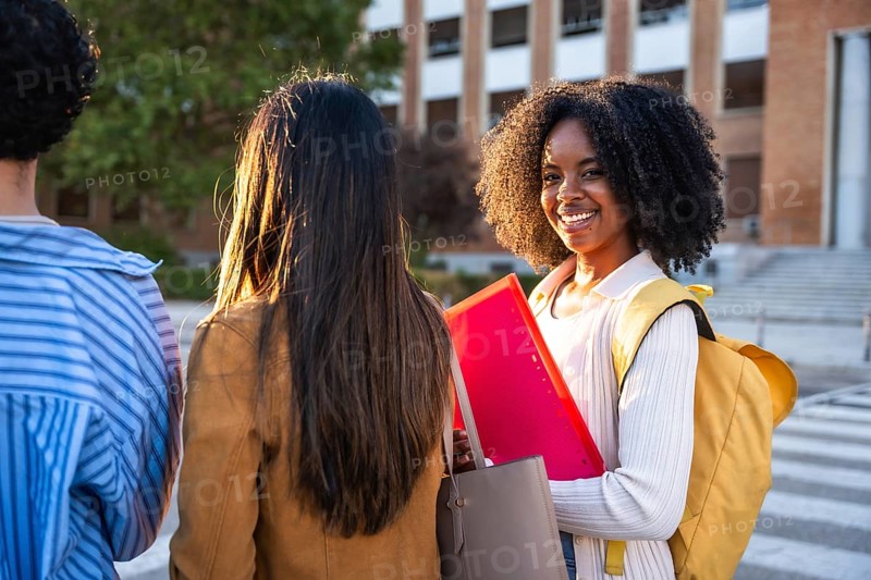 Group of young diverse women students walking together, with one black woman looking over shoulder and smiling warmly while carrying books and backpack on campus