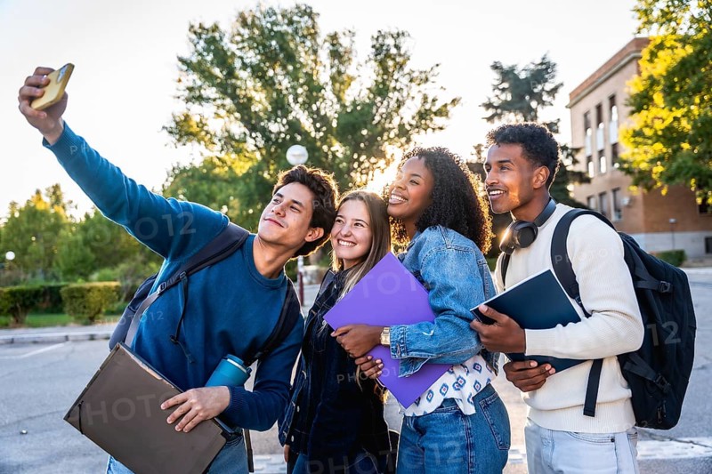 Diverse group of smiling university students recording a selfie outdoors on campus, holding books and backpacks, representing friendship, education, and youth