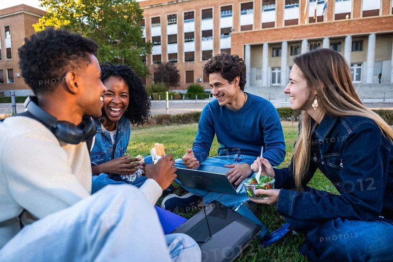 Group of happy young students smiling and laughing, sitting on grass having lunch and working with a laptop in front of a university building on a sunny day