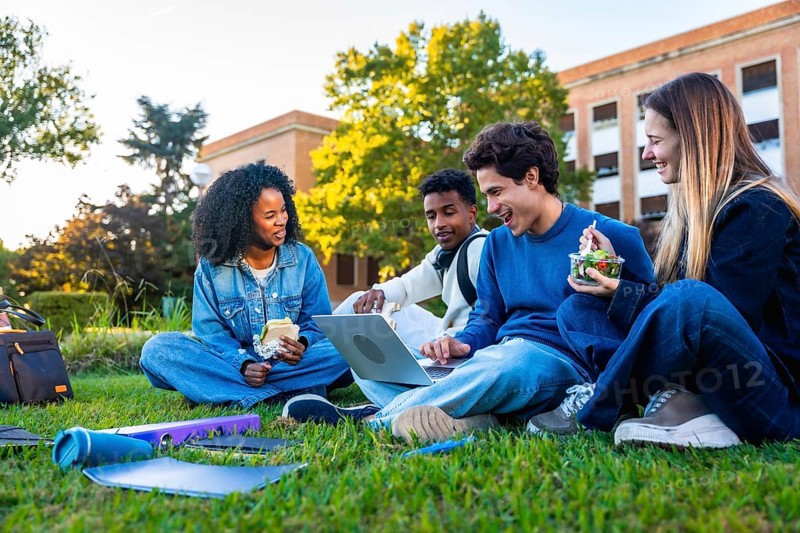 Group of diverse university students sitting on the green grass campus, sharing a laptop, studying, smiling, and enjoying lunch together during a break