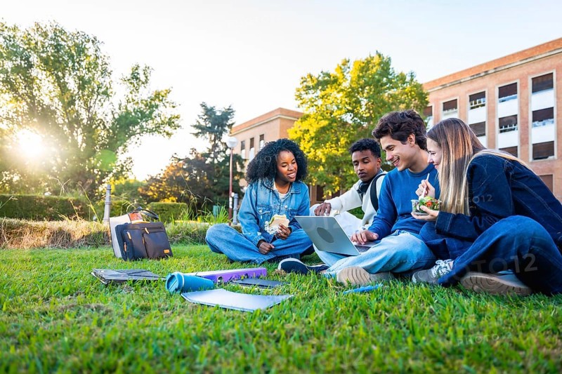 Group of multiethnic university students sitting on green grass, studying and discussing coursework on a laptop, enjoying a break with food and drinks on campus during a sunny day