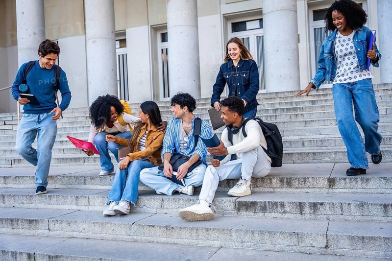 Diverse young students chatting, laughing and walking on wide stone campus steps by a university building, enjoying a casual outdoor study break and friendly conversation