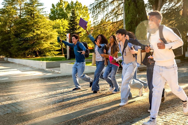 Group of excited multiracial students. Wearing casual clothes and carrying backpacks and books. Running across a university campus with trees in the background. Celebrating success or rushing to class