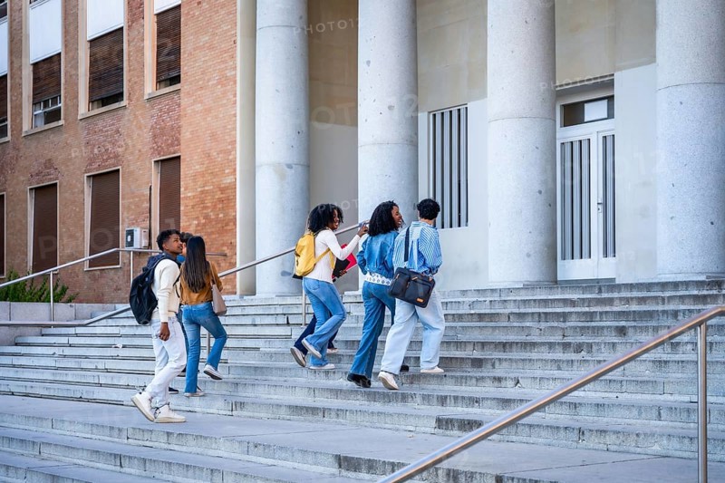 Diverse group of college students walking up wide outdoor steps toward a university building with large columns, backpacks and books, heading to class or campus event
