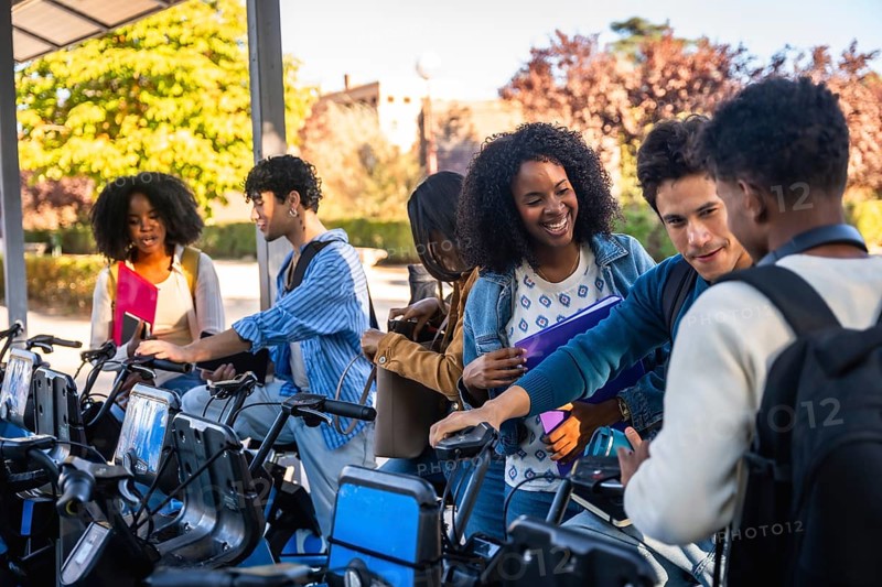Group of happy diverse university students activating a shared bicycle from a station, preparing to ride on campus, representing sustainable urban transport
