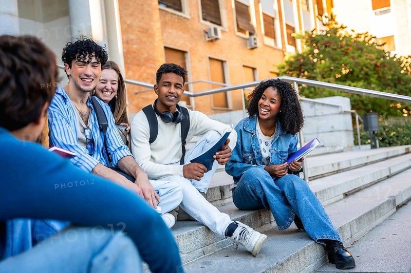 Diverse group of multiracial youth sitting on campus steps, engaging in conversation and smiling warmly, embodying friendship and education outside a university building