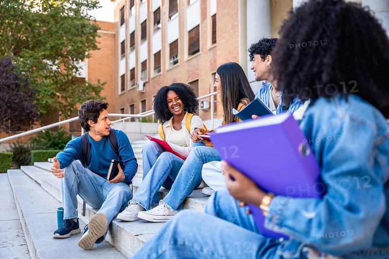 Diverse group of university students sitting on campus steps, smiling and chatting confidently, sharing books and backpacks while enjoying outdoor study, friendship and learning