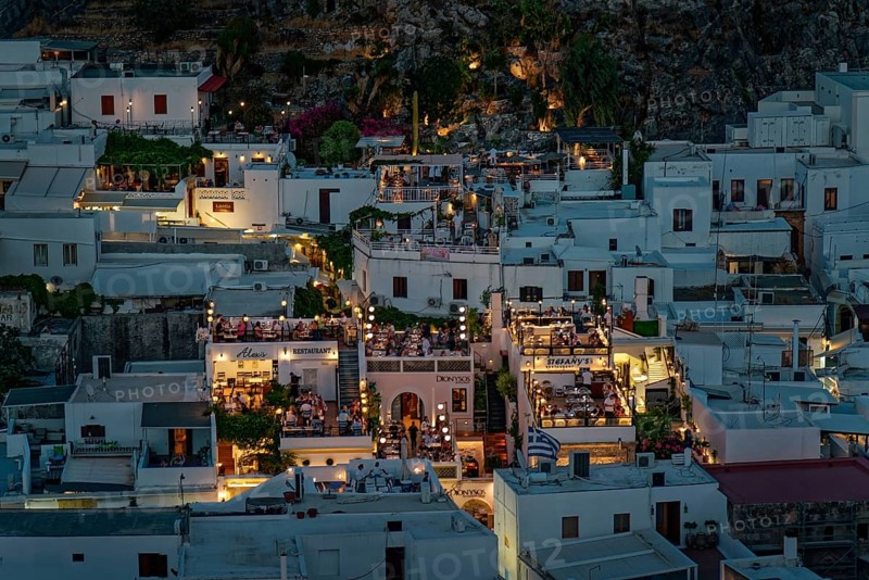 View of the illuminated roof terraces and restaurants of the town of Lindos and white houses that radiate a cosy atmosphere, Acropolis, Lindos, Rhodes, Island, Greece