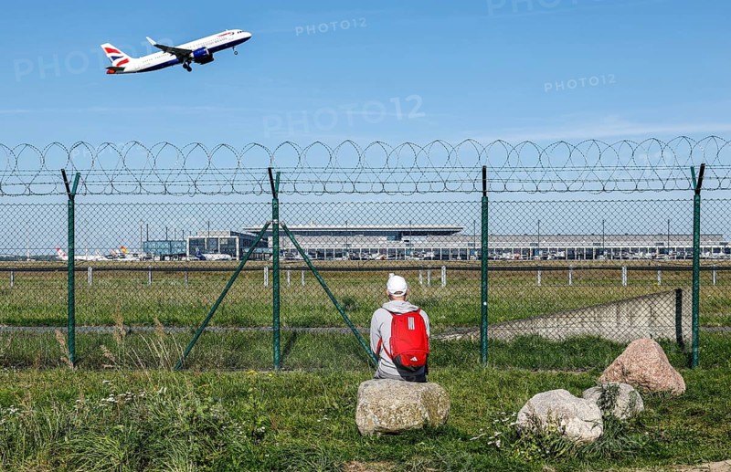 A man sits on a stone and watches the take-off of a British Airways plane through a barbed wire fence at BER Airport, Berlin Schönefeld, 28/09/2025, Schönefeld, Brandenburg, Germany