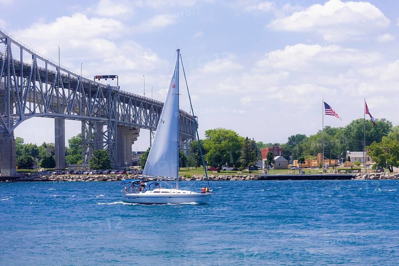 Sailboat gliding near the Blue Water Bridge, connecting Canada and the United States, with flags under a bright blue sky along the St. Clair River leading to Lake Huron in Sarnia, Ontario, Canada, North America