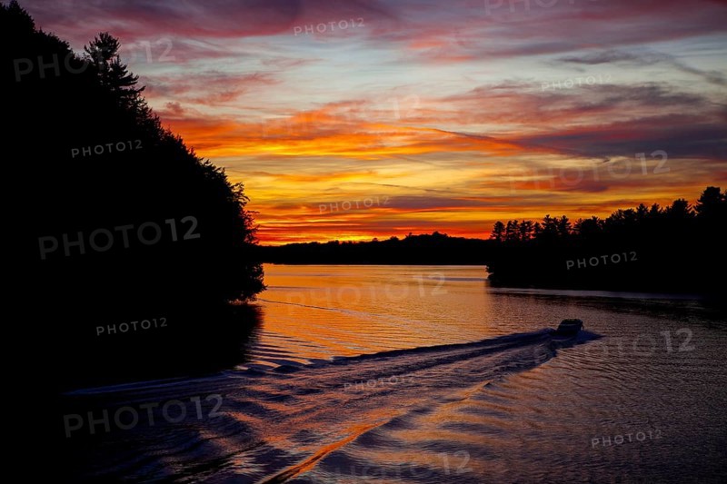 A boat creates ripples in a lake at sunset, with vibrant colors and silhouetted trees in Muskoka, Ontario, Canada, North America