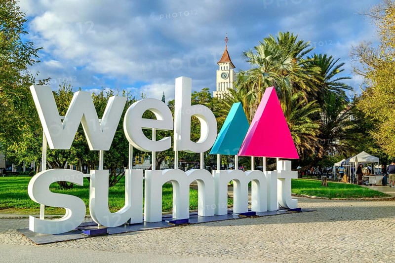 Nov 13, 2024 - Lisbon, Portugal: Colorful letters spell out Web Summit, influential technology conference, near park with lush palm trees and historic clock tower on sunny day. Discussions, network, innovative ideas, dynamic atmosphere of event