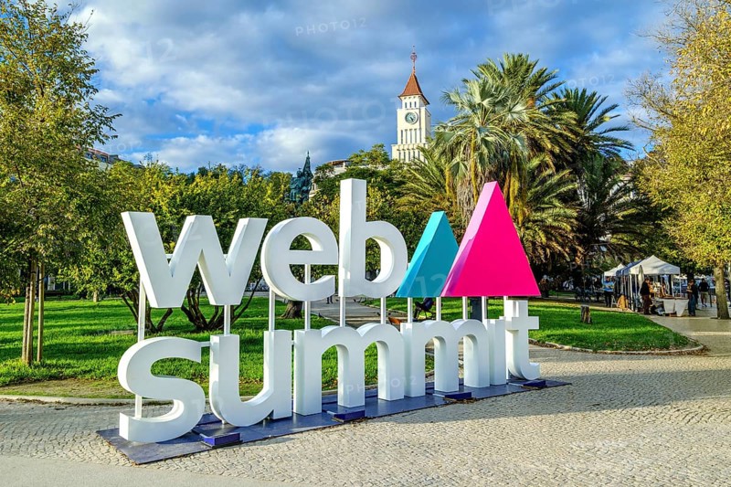 Nov 13, 2024 - Lisbon, Portugal: Large sign displaying Web Summit logo in lively park filled with greenery, historic tower in background. Major annual technology conference internet technology, emerging technology, marketing, AI solution, networking