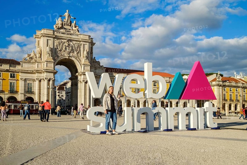 Nov 13, 2024 - Lisbon, Portugal: A female adult posing for a photo in front of Web Summit sign installation at Commerce square and Rua Augusta Arch. People walking, international technology conference, discussions, networking, technology, innovation