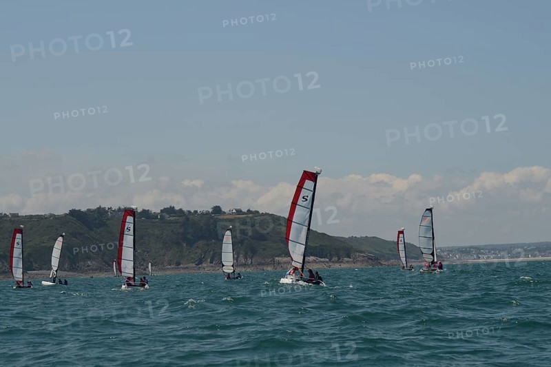 Sailboats glide on the ocean near a hilly coastline under a clear sky, in Brittany, France