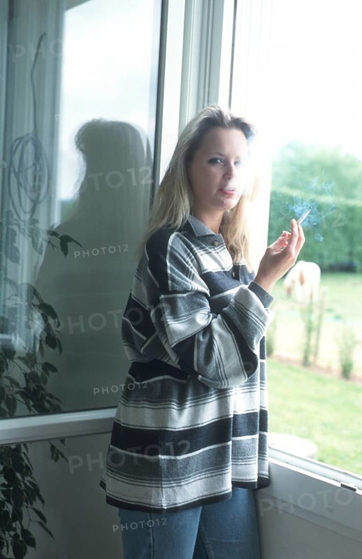 A woman with a striped shirt stands by a window, holding a cigarette, with natural light illuminating her