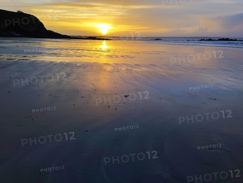 A serene sunset over a beach with calm ocean waters and rocks silhouetted against a golden sky