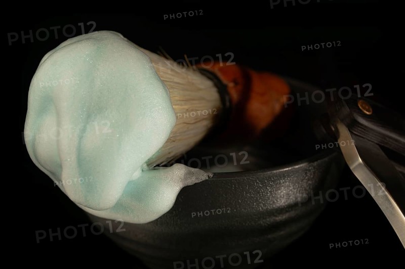 Shaving brush with teal lathered foam and straight razor in a dark bowl for men's grooming
