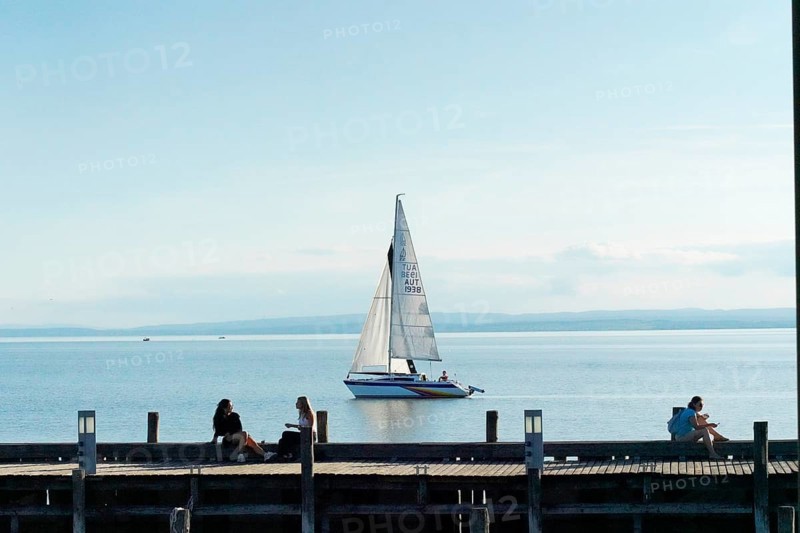 Lake Neusiedl, Burgenland, Austria, Europe, A sailboat sails calmly on a lake while people relax on the jetty, Sailboats on Lake Neusiedl