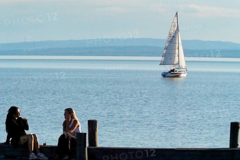 Lake Neusiedl, Burgenland, Austria, Europe, Two woman sit on the jetty while a sailing boat passes by on the calm lake, Sailing boats on Lake Neusiedl