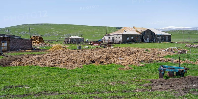 A farm with old buildings and agricultural machinery on a green pasture, cow dung in front of it, Berdashen, Lake Arpi, Shirak province, Armenia