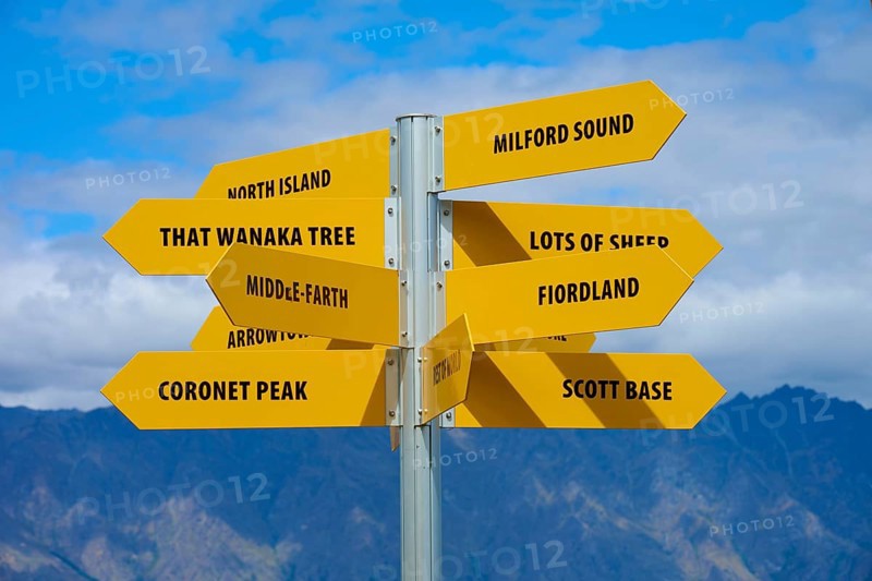 Yellow signpost with several signs and a mountain ridge in the background against a blue sky, Queenstown, Queenstown-Lakes District, Otago, South Island, New Zealand