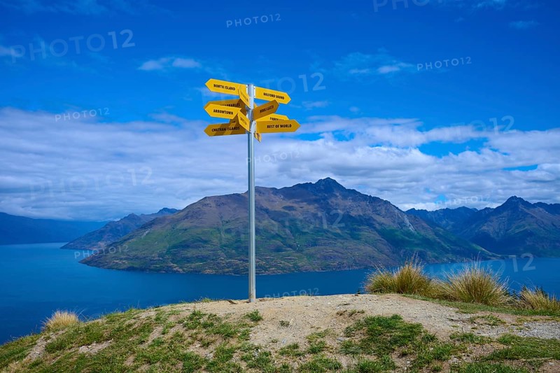 Yellow signposts on a hill in front of a vast mountain landscape under a blue sky, Queenstown, Queenstown-Lakes District, Otago, South Island, New Zealand