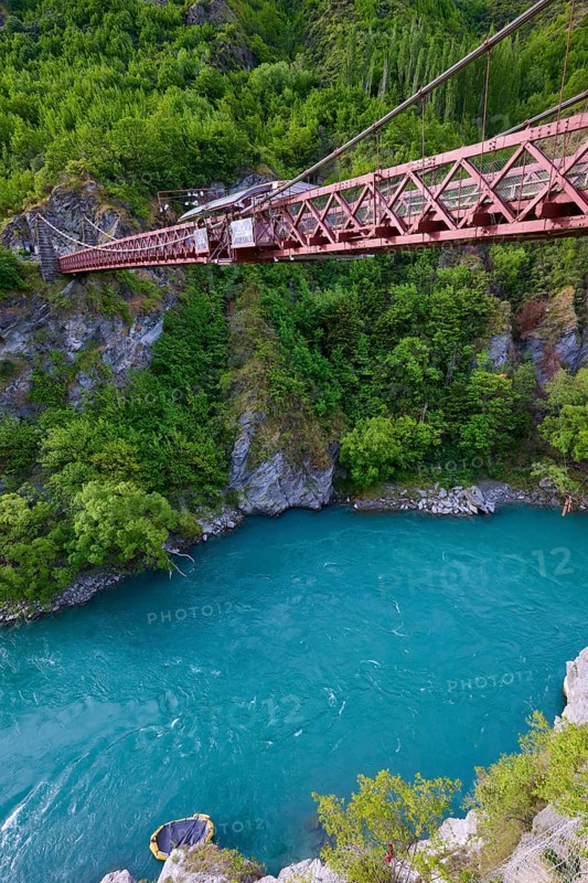 Red suspension bridge over river with steep wooded cliffs and calm waters, Kawarau Gorge Suspension Bridge, Kawarau River, Queenstown, Otago, South Island, New Zealand