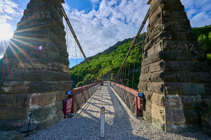 Old bridge with massive stone pillars surrounded by lush greenery under a bright blue sky, Kawarau Gorge Suspension Bridge, Kawarau River, Queenstown, Otago, South Island, New Zealand